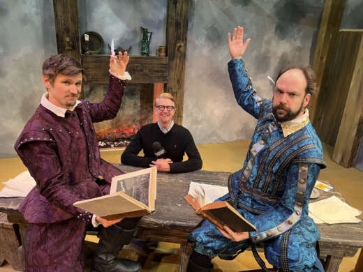 Wild Bill Wood sits behind a table on set at Le Petit Theatre. In front are actors in costume: Dylan Hunter as William Shakespeare and Ian Hoch as Christopher "Kit" Marlowe. Both actors hold open books and have one arm raised above their heads.