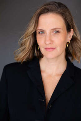 Julie Lake is shown in front of a gray backdrop. She smiles lightly into the camera, and is wearing a black blouse with deep V neckline and gold drop-style earrings.